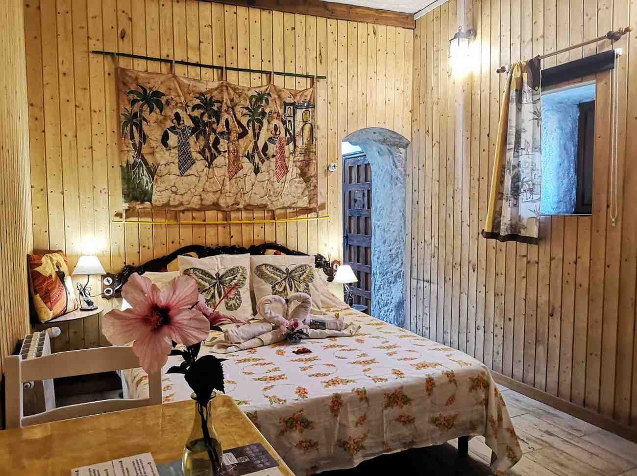 Dormitorio con paredes de madera, cama con ropa de cama de flores y tapiz colorido. Hay una pequeña ventana a la derecha.