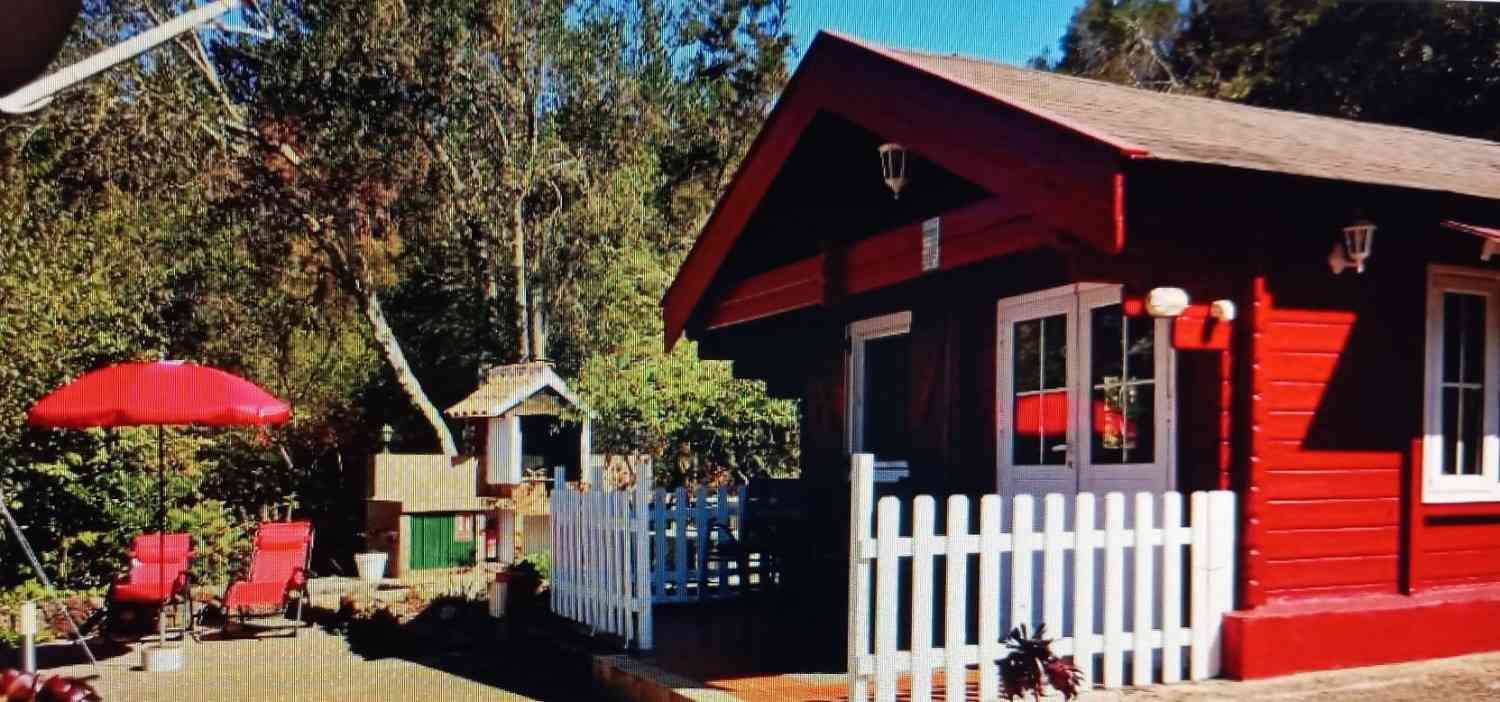 Cabaña roja con cerca blanca, sillas, sombrilla y una casa de juegos en un patio verde.