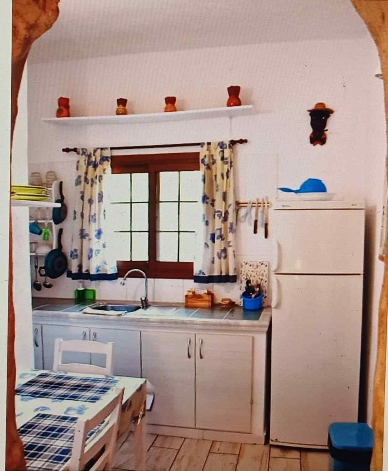 Acogedora cocina blanca con detalles en azul. Ventana pequeña, refrigerador y mesa. Estante decorativo con cerámica.