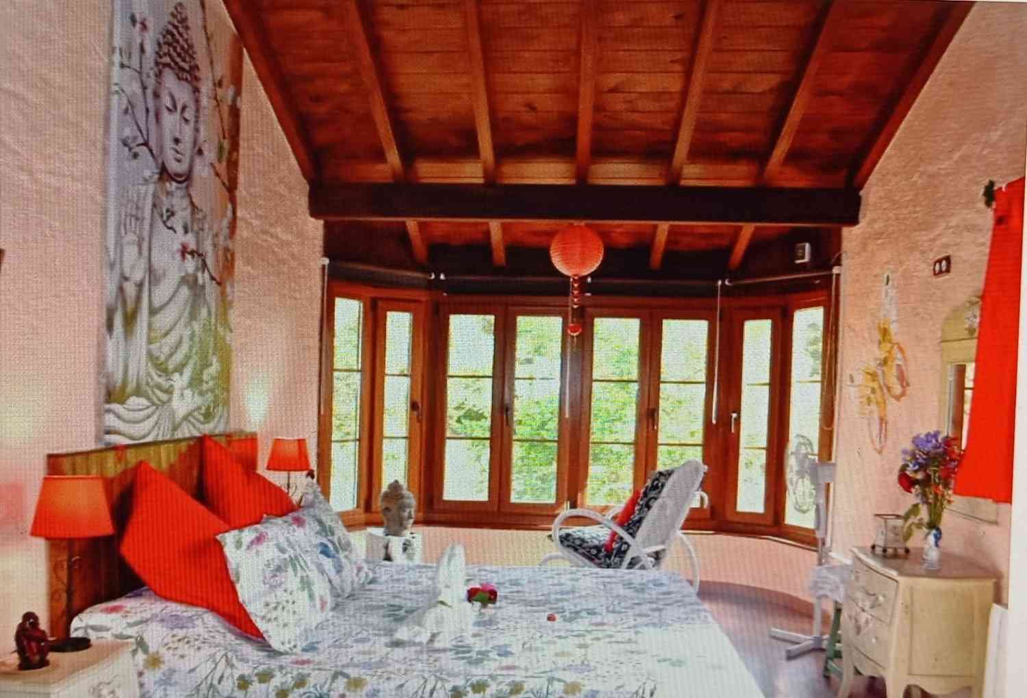 Dormitorio con ventanal, techo de madera, tapiz de Buda, almohadas rojas y un sillón.