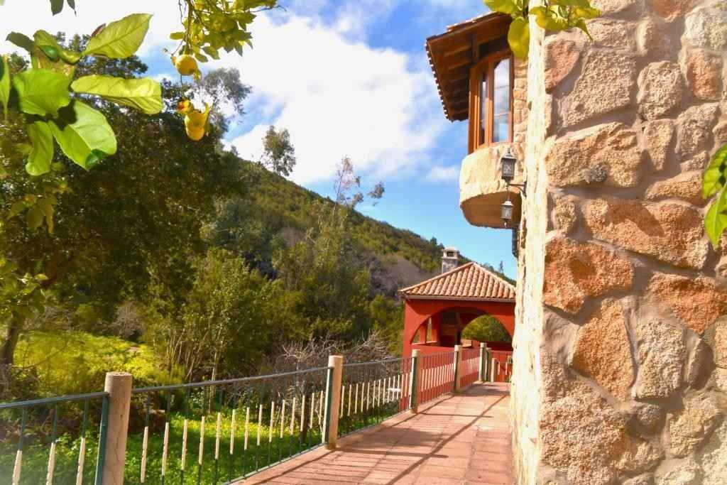 Edificio de piedra con balcón y pasarela junto a una ladera verde y limonero.