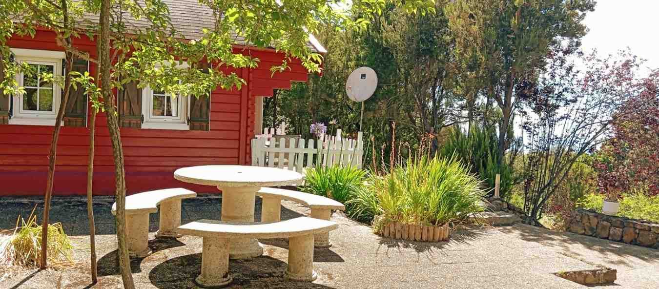 Casa de juegos roja y mesa de picnic en un jardín con árboles y valla blanca.
