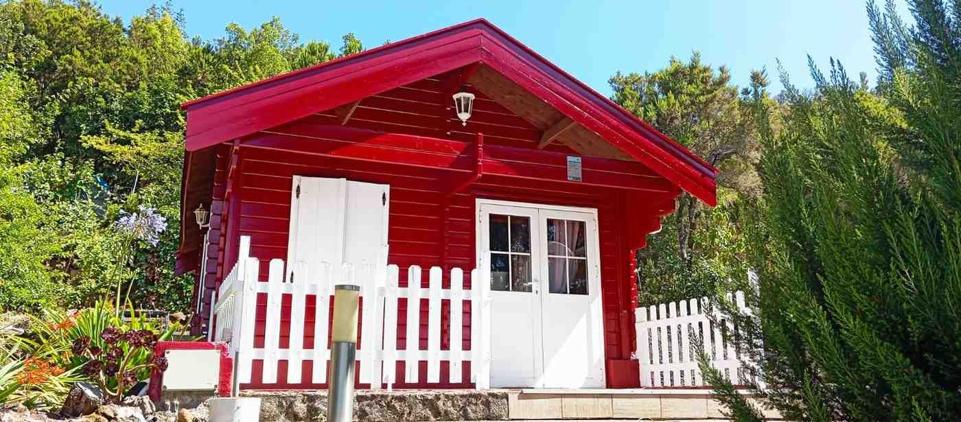 Cabaña roja con valla blanca, puertas y contraventanas rodeadas de follaje verde.