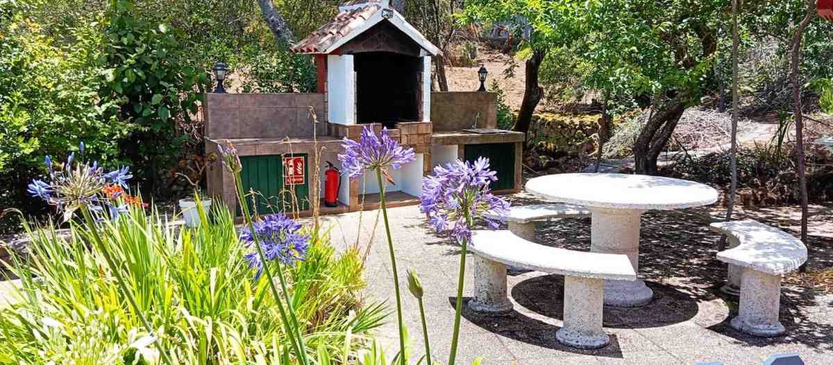 Mesa de picnic de piedra con bancos, flores violetas y un pequeño edificio en un entorno de jardín.