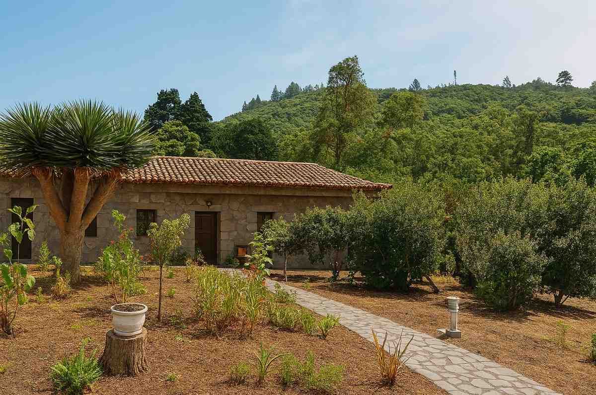 Casa de piedra con tejado de tejas, rodeada de vegetación y con una montaña al fondo. Un camino de piedra conduce a la casa.