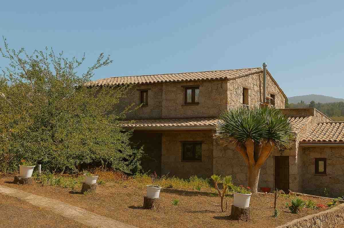 Casa de piedra con techo de tejas, rodeada de árboles y un paisaje marrón bajo un cielo despejado.