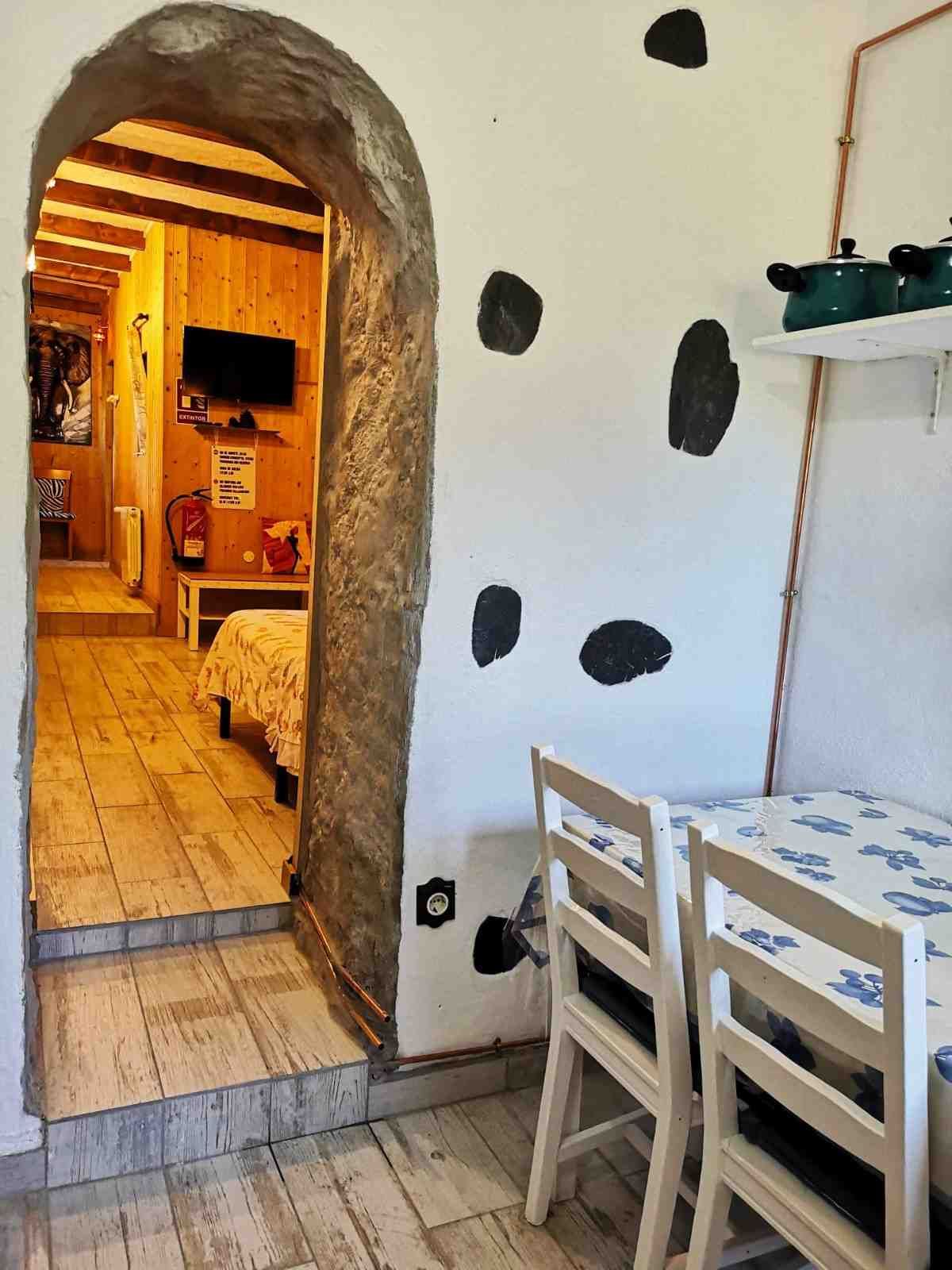 Vista interior con arco de piedra que da acceso a un dormitorio. Paredes blancas con manchas oscuras, mesa y sillas.