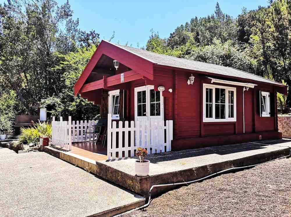 Cabaña roja con detalles blancos y cerca de estacas, ubicada en una zona boscosa bajo un cielo azul brillante.