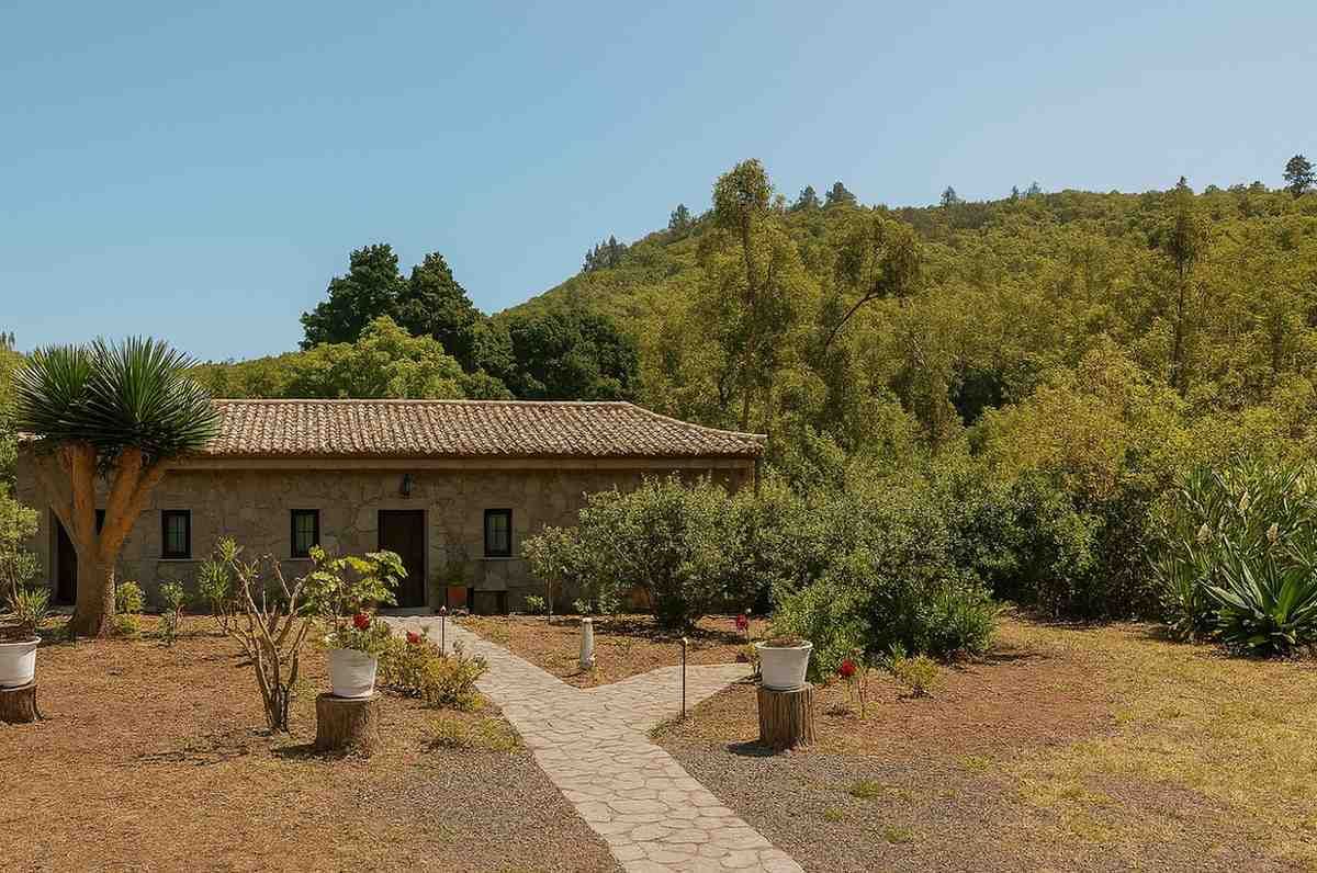 Casa de piedra con tejado de teja, rodeada de árboles y camino de piedra, situada contra una ladera.