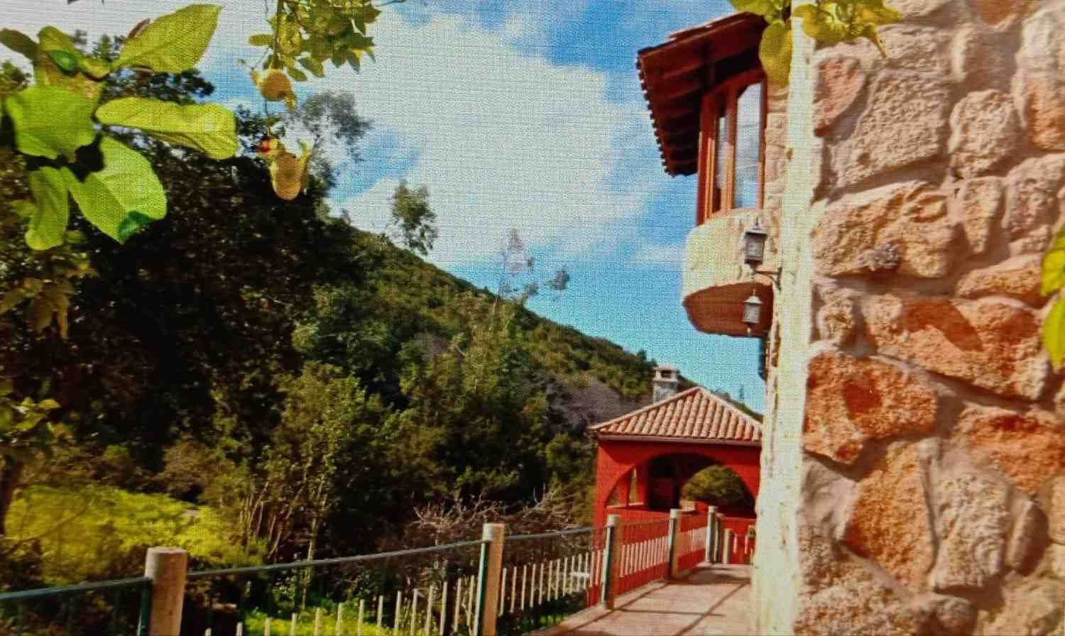 Edificio de piedra con detalles en rojo, balcón, pasarela y exuberante ladera.