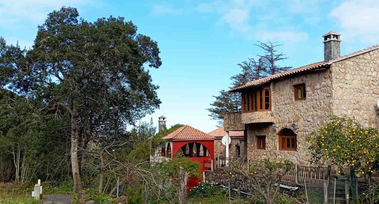 Casa de piedra con detalles en rojo, rodeada de árboles y vegetación, contra un cielo azul.