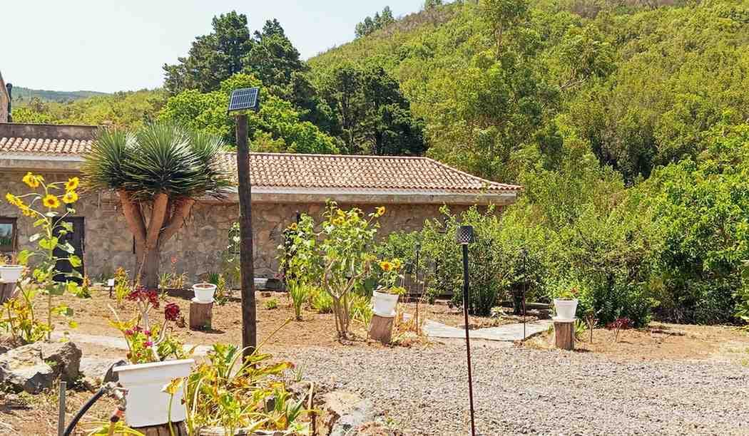 Edificio de piedra con techo de tejas, rodeado de jardín, árboles y ladera en un día soleado.