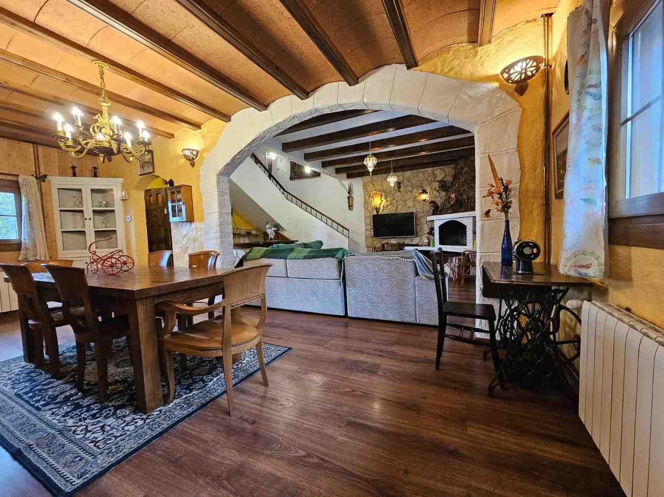 Comedor con mesa de madera, alfombra, arco de acceso a salón con chimenea y escalera.