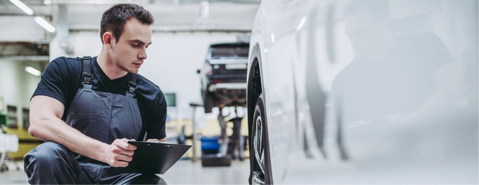 Ein Mann kniet in einer Garage vor einem Auto und schaut auf ein Klemmbrett.