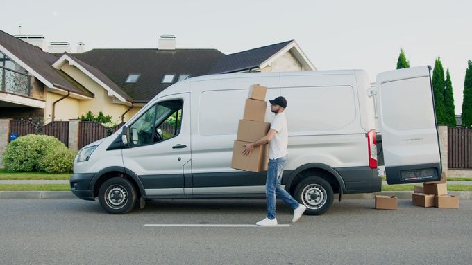 Un hombre lleva cajas delante de una camioneta blanca.