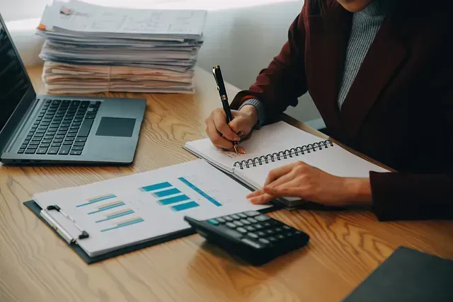 Una persona con una chaqueta de traje color burdeos trabaja en un escritorio, escribiendo en un cuaderno con un bolígrafo, revisando gráficos y usando una calculadora y una computadora portátil cerca.