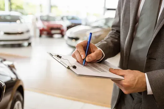 Hombre de traje escribiendo en un portapapeles en un concesionario de automóviles, con varios coches en el fondo.