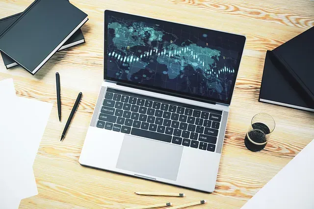 Computadora portátil que muestra un mapa del mundo y datos financieros en un escritorio de madera con libros, bolígrafos y papeles.