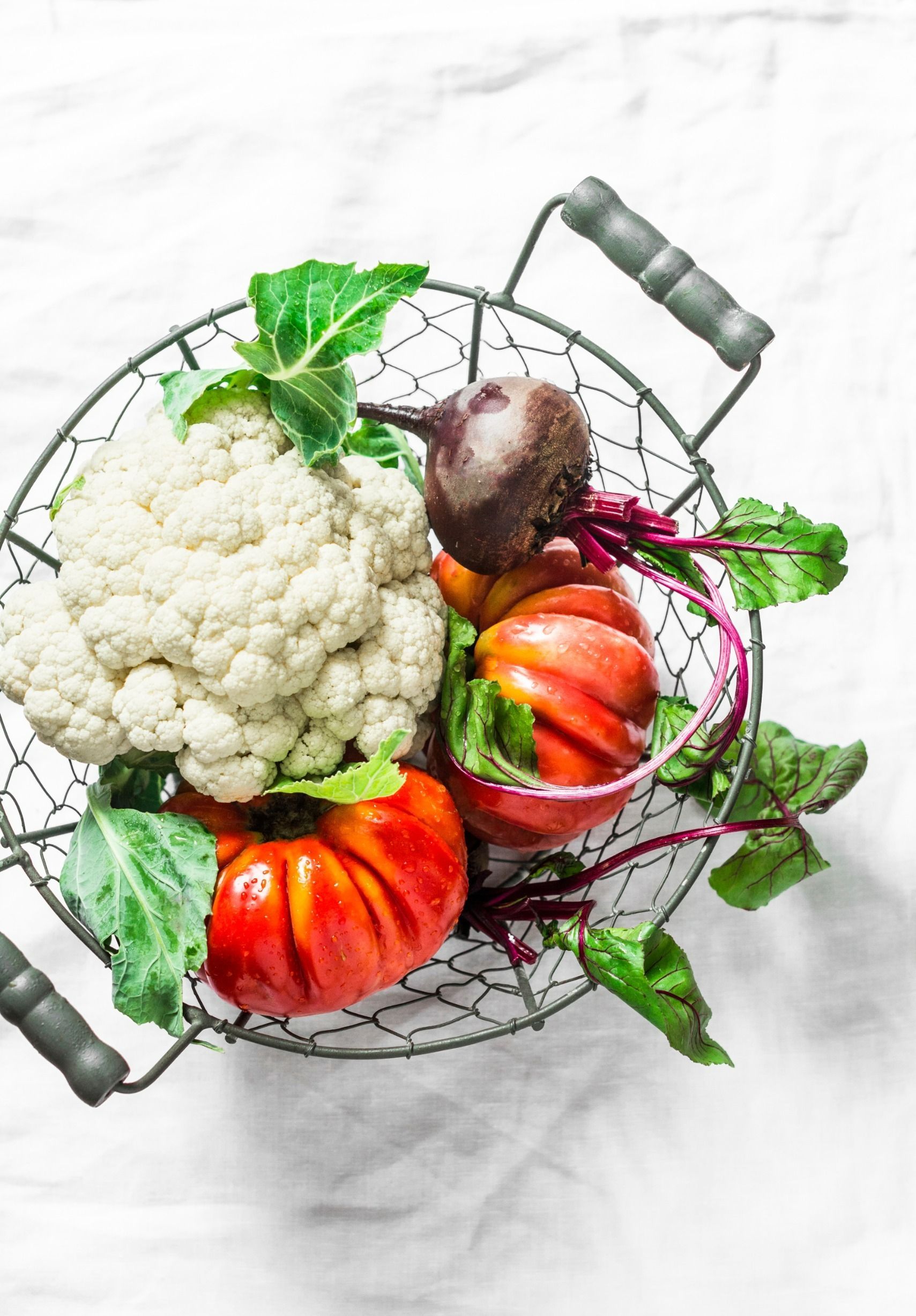 Una cesta de alambre llena de coliflor, remolacha, tomates y otras verduras.
