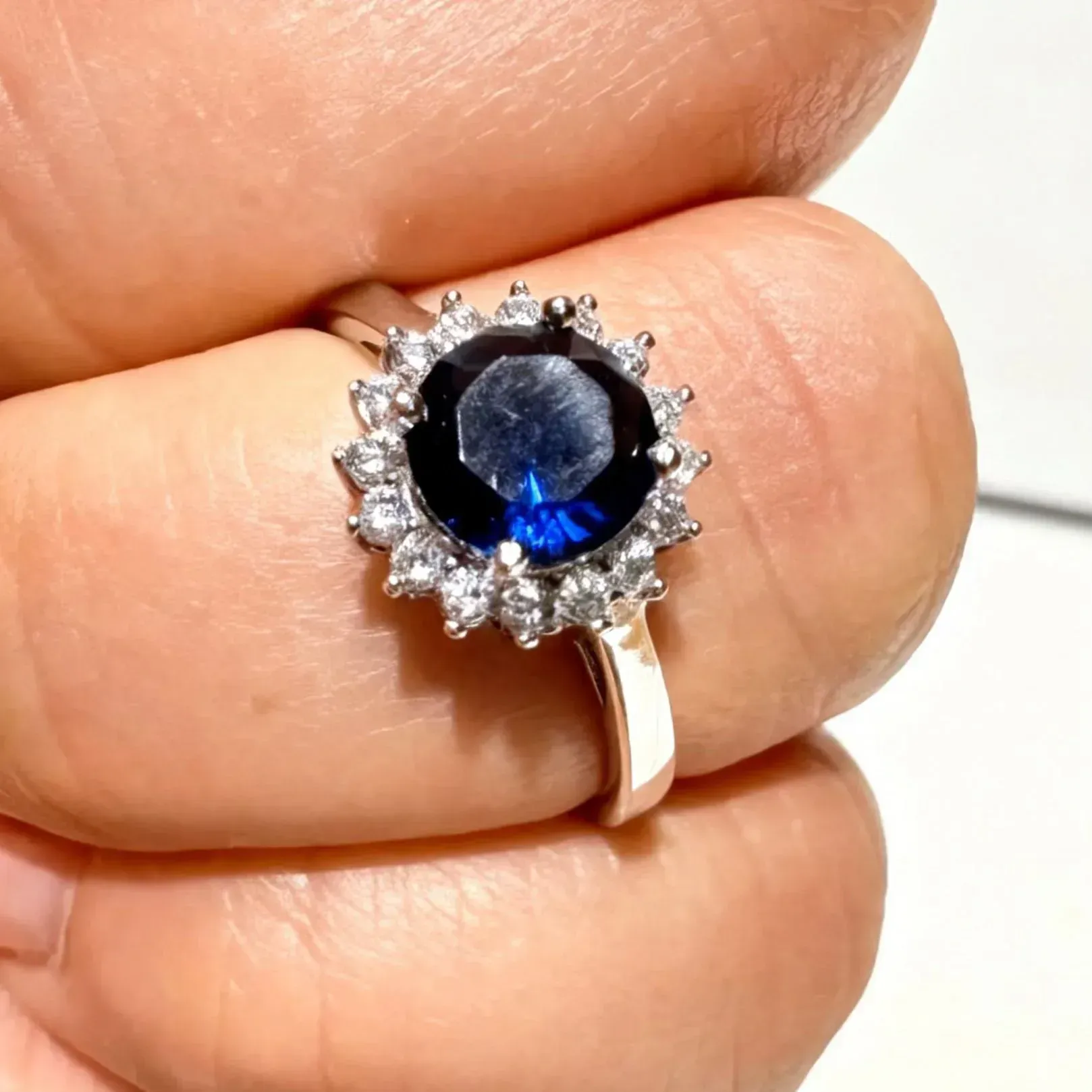 Anillo de plata con una gema azul redonda y piedras transparentes en forma de halo en los dedos.