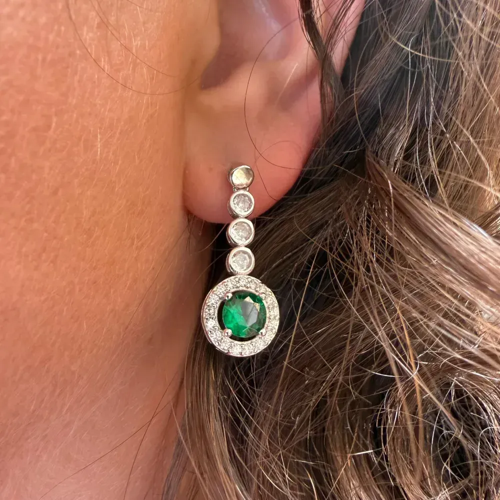 Primer plano de un pendiente colgante de plata con piedras preciosas transparentes y verdes sobre el cabello y la piel.