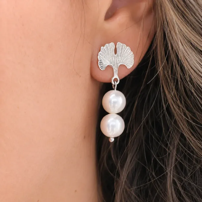 Primer plano de un pendiente plateado en forma de hoja con dos perlas blancas en una oreja.