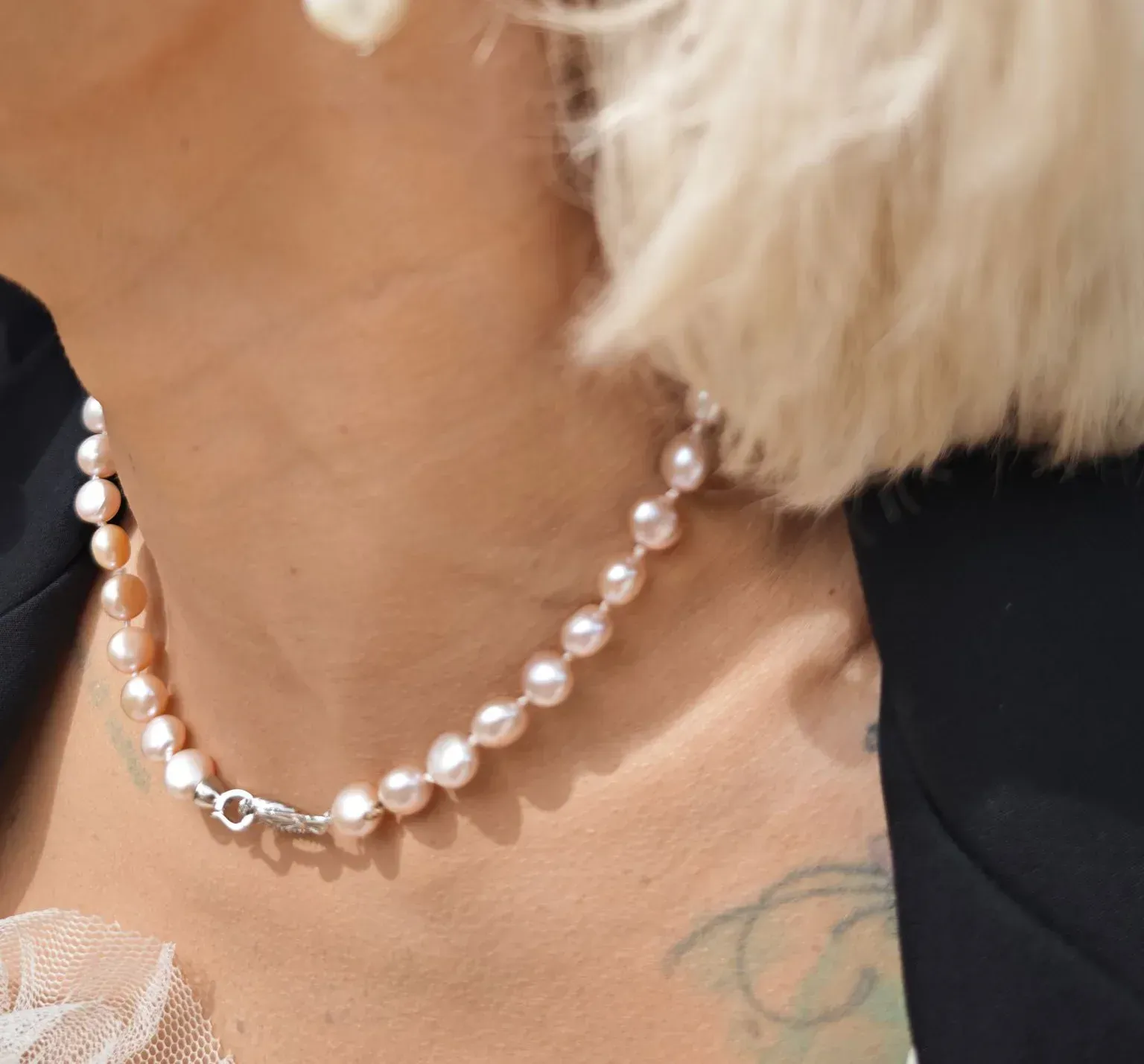 Primer plano de un collar de perlas en el cuello, con cabello rubio corto y un blazer negro visibles.