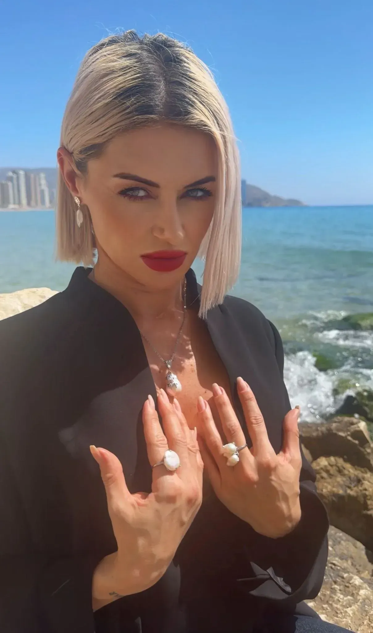 Una persona posando en una playa soleada, con una chaqueta negra y pintalabios rojo.