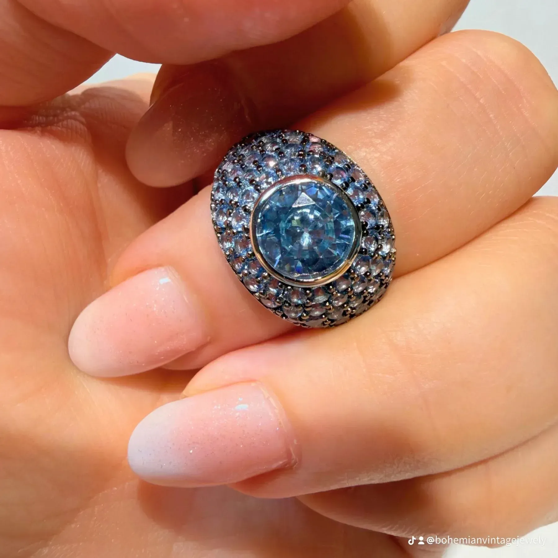 Primer plano de una mano sosteniendo un anillo con una piedra preciosa azul rodeada por un halo de pequeños diamantes.