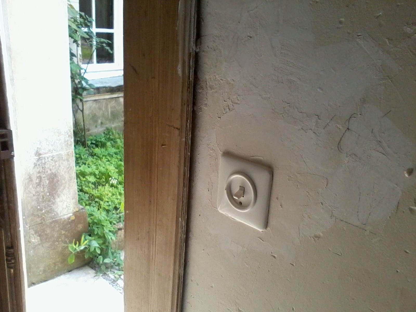 Mur blanc avec un petit interrupteur carré à côté d'une porte ouverte donnant sur l'extérieur.