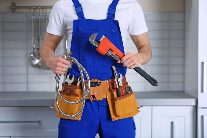 Personne en salopette bleue tenant une clé et une perceuse orange, portant une ceinture à outils dans un décor de cuisine.