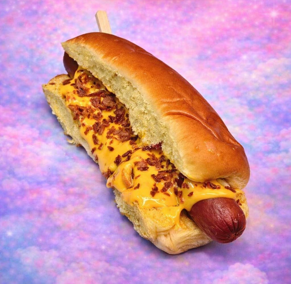 Perrito caliente en un panecillo con mostaza y queso, cubierto con trocitos de tocino, sobre un fondo rosa-morado.