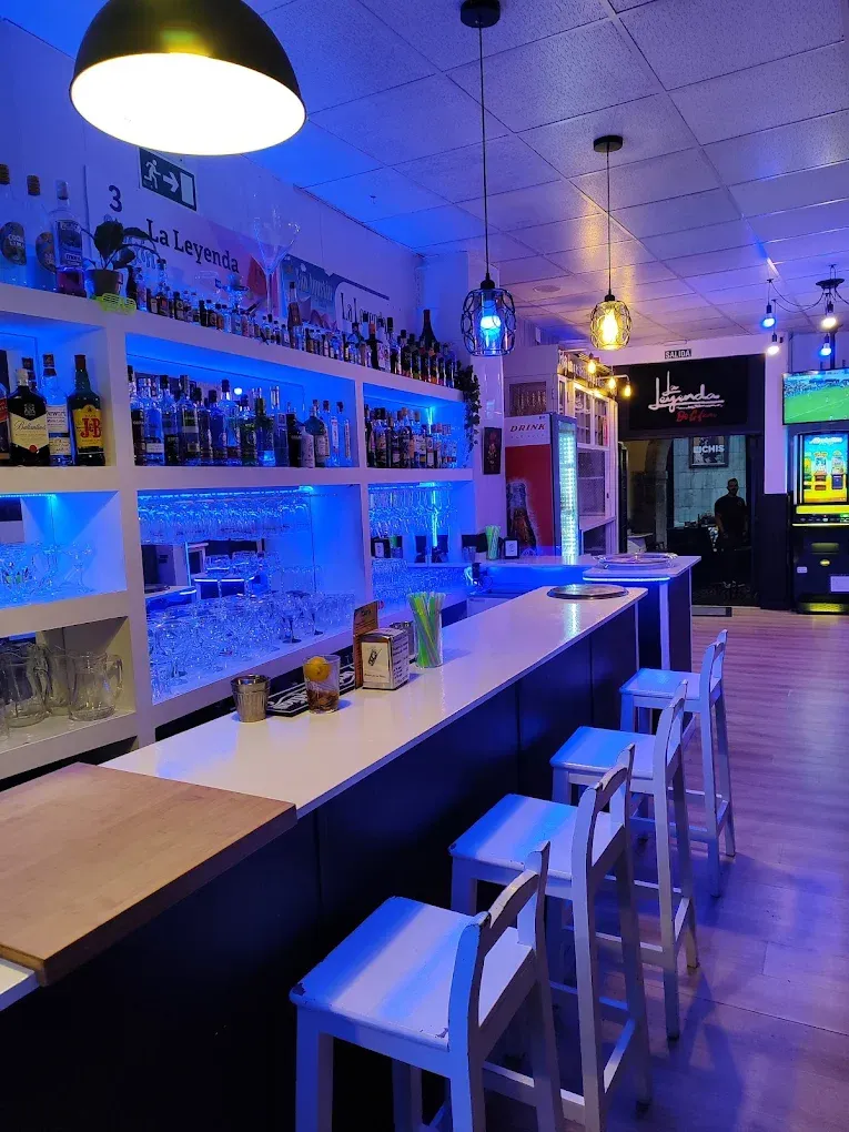 Interior de bar moderno con iluminación azul, taburetes, estantes para botellas y lámparas colgantes.