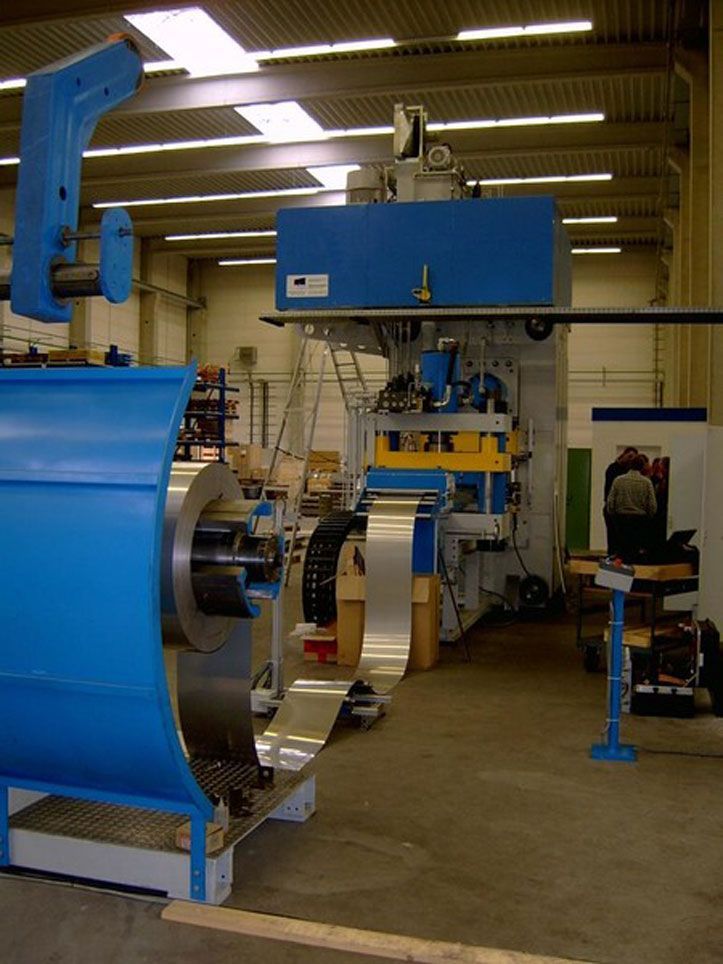 In einer Fabrik wird eine Metallspule in eine Stanzmaschine abgewickelt. Blaue und silberne Maschinen.