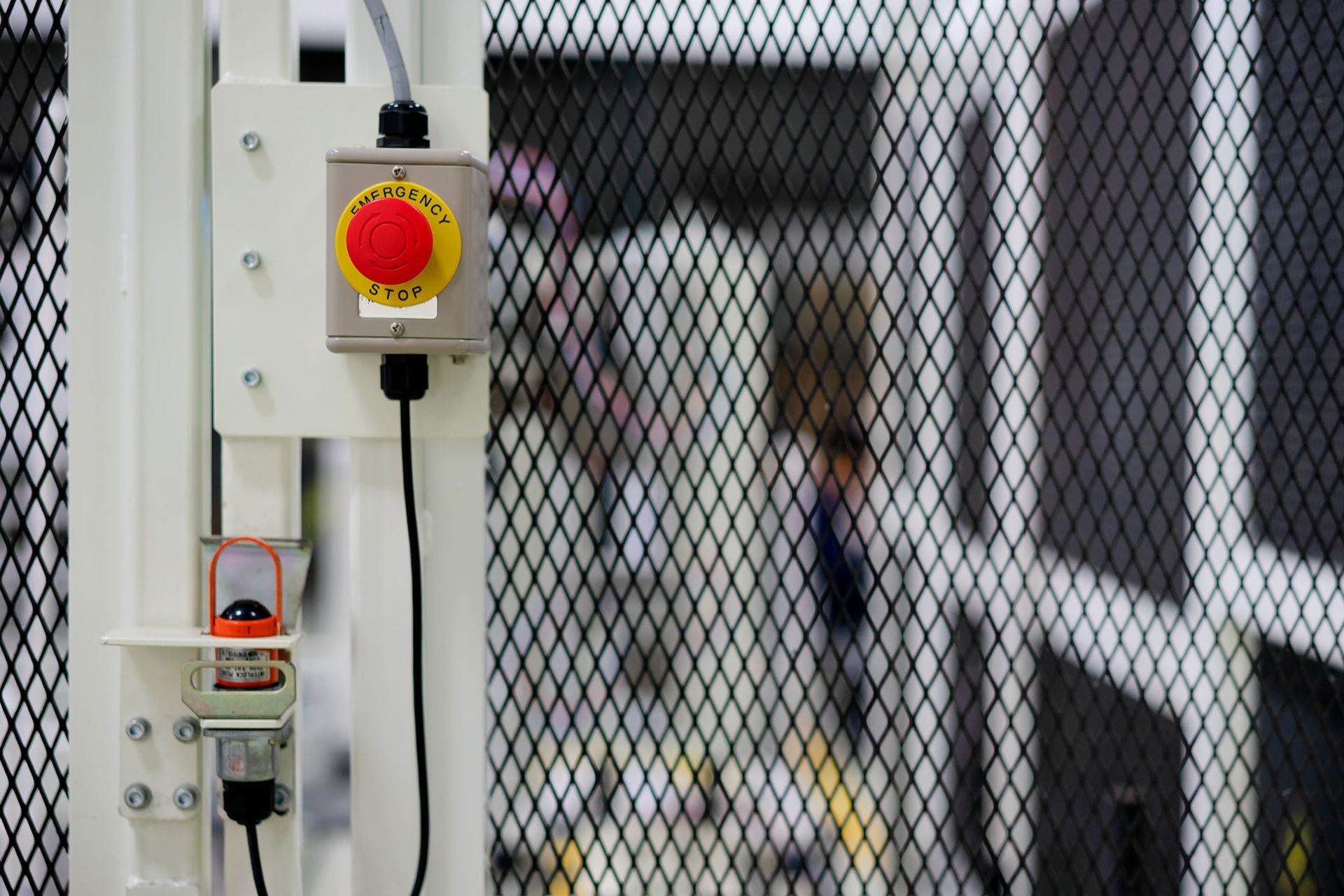 Roter Not-Aus-Knopf auf weißem Bedienfeld, neben einer schwarzen Drahtgitter-Sicherheitsbarriere.