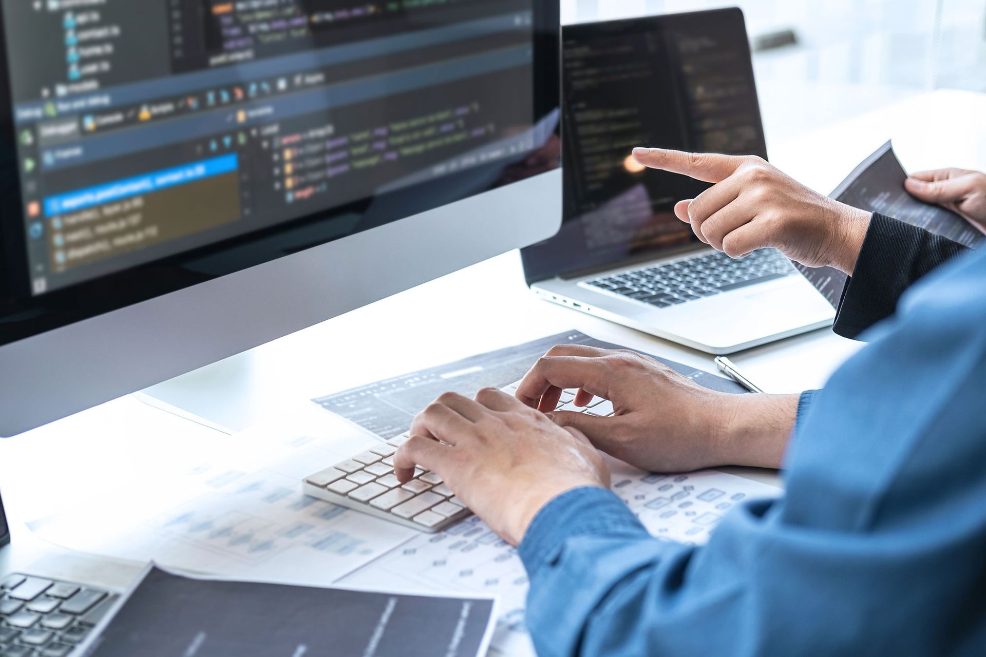 Zwei Personen arbeiten an Computern, eine tippt, die andere zeigt auf einen Bildschirm, auf dem Code angezeigt wird.