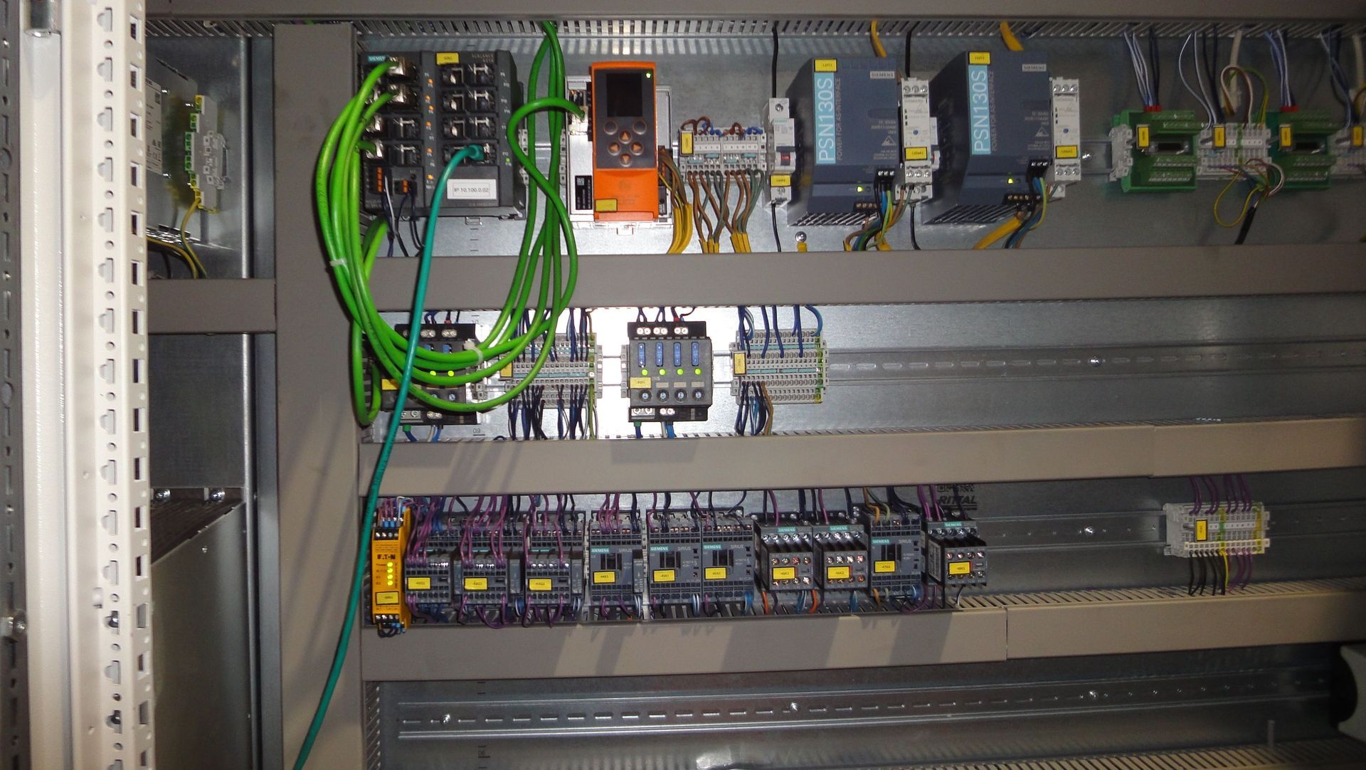 Elektrischer Schaltschrank mit verschiedenen Bauteilen, grünen und schwarzen Drähten und grauer Metallrückwand.