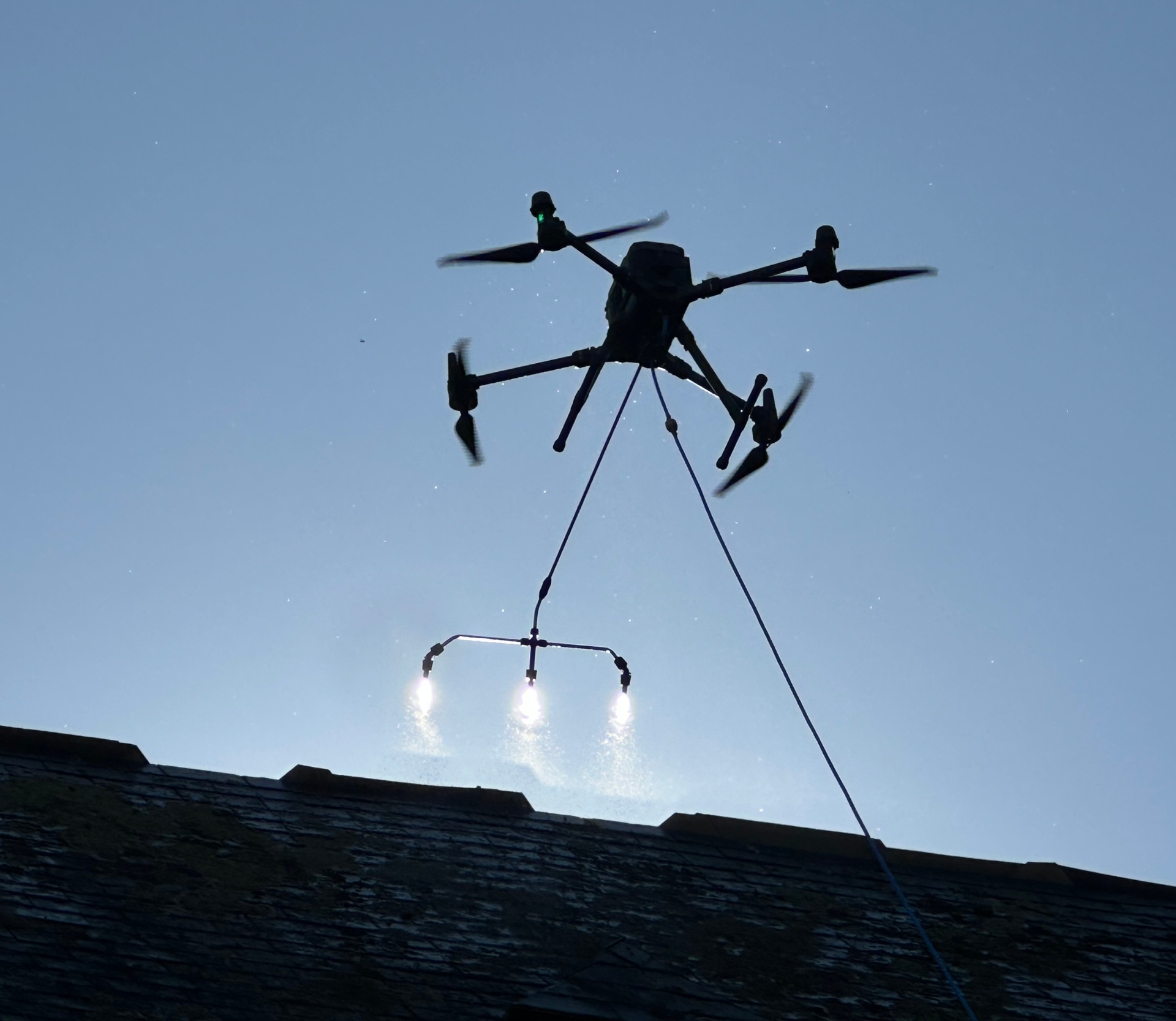 Un drone survole un toit, l'illuminant grâce à ses projecteurs. Sa silhouette se détache sur un ciel lumineux.