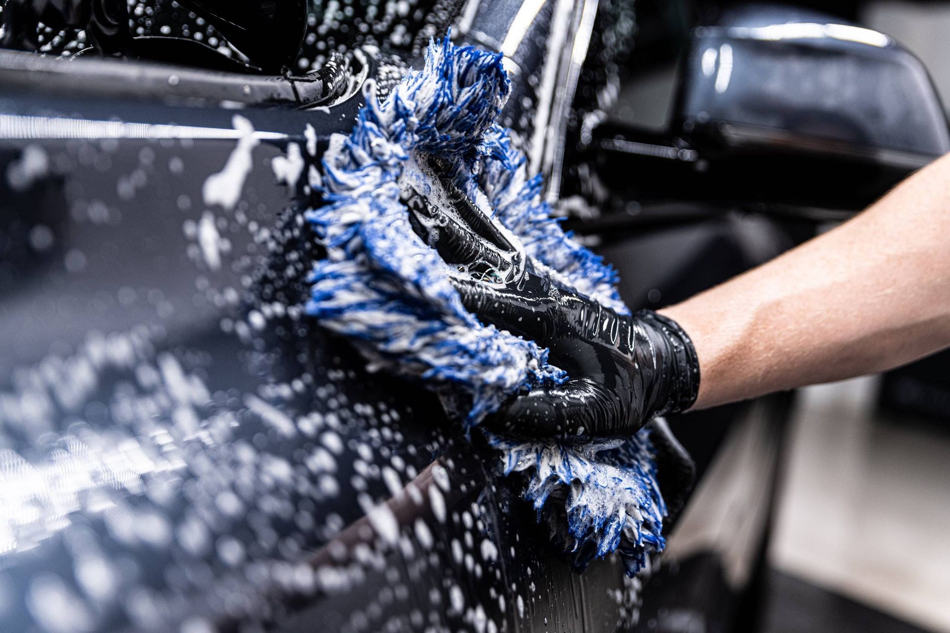 Main gantée lavant une voiture noire avec un gant en microfibre bleu, de l'eau éclaboussant la carrosserie