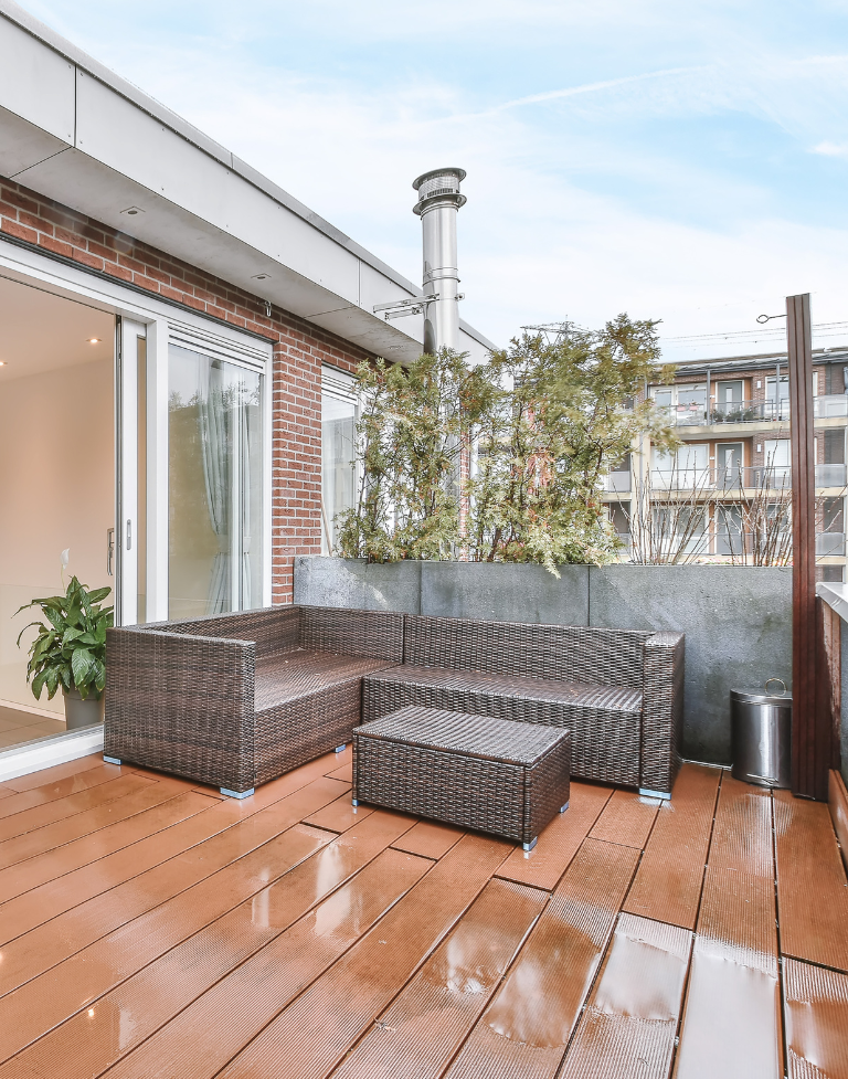 Terrasse extérieure avec mobilier en osier et parquet. Porte coulissante en verre, mur de briques et végétation abondante.