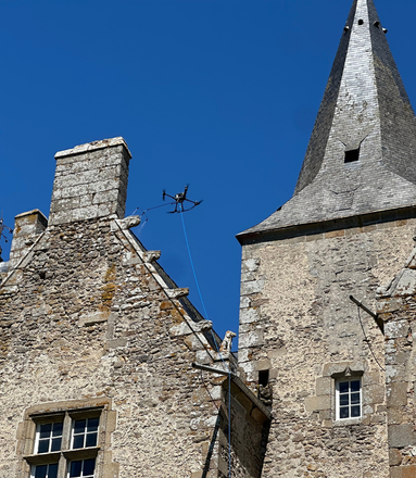 Un drone inspecte la cheminée et la tourelle d'un bâtiment en pierre sous un ciel bleu clair.
