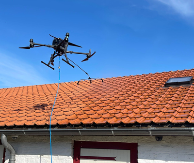 Un drone nettoie un toit en tuiles rouges avec un tuyau bleu contre un ciel bleu vif.