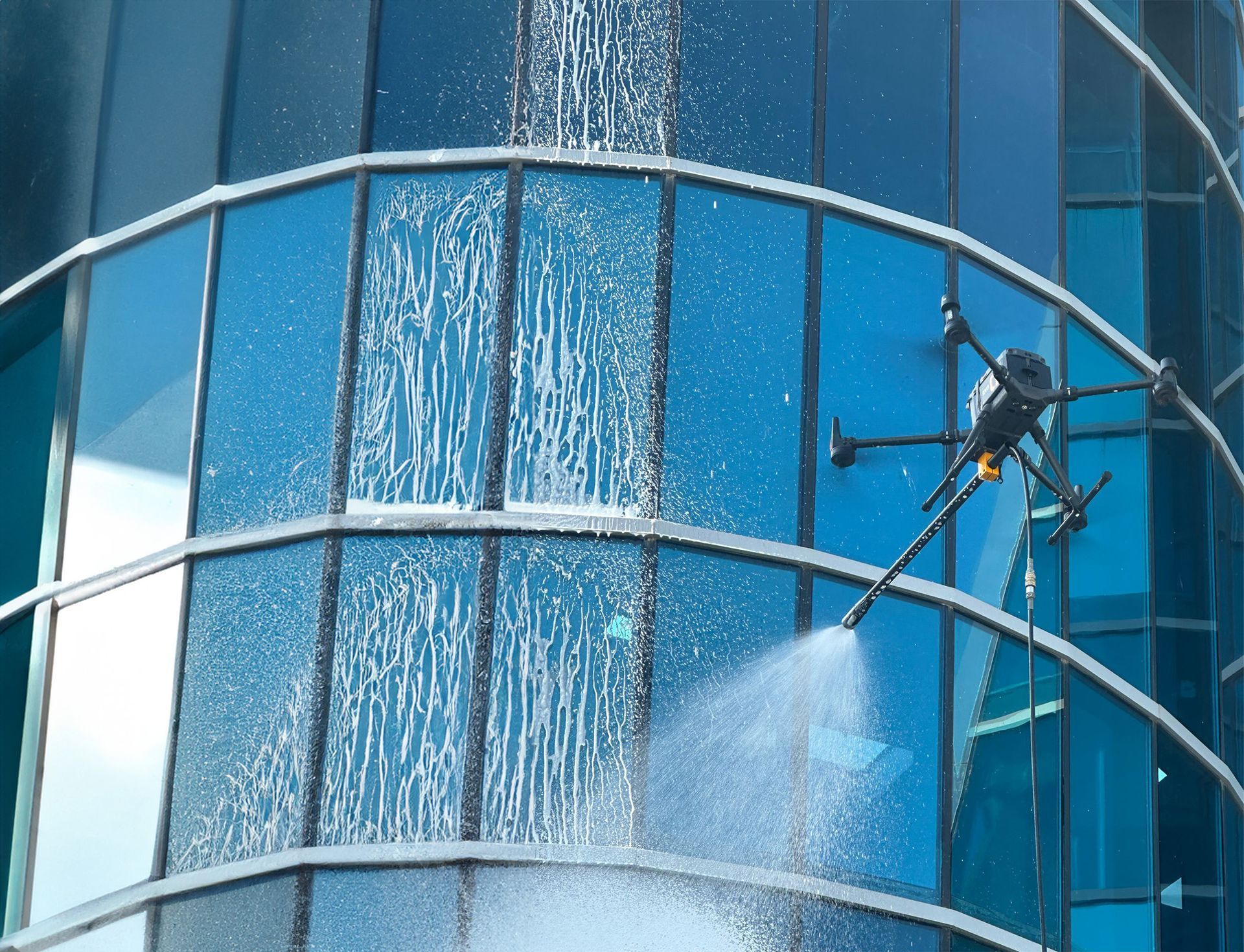 Un ouvrier nettoie à haute pression un bâtiment en verre bleu depuis une nacelle suspendue.
