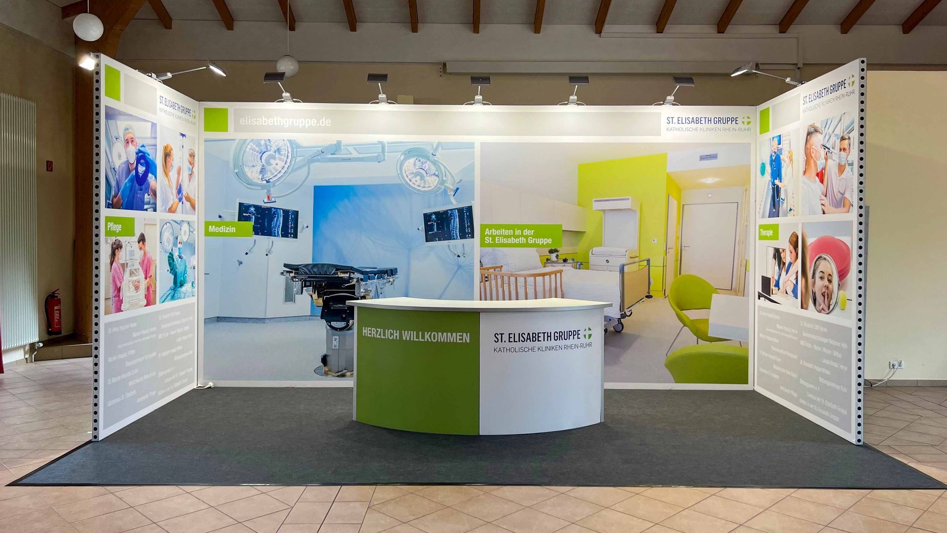 Messestand mit weißen Paneelen, grünen Akzenten, Ausstellungsplakaten und einem zentralen Tresen