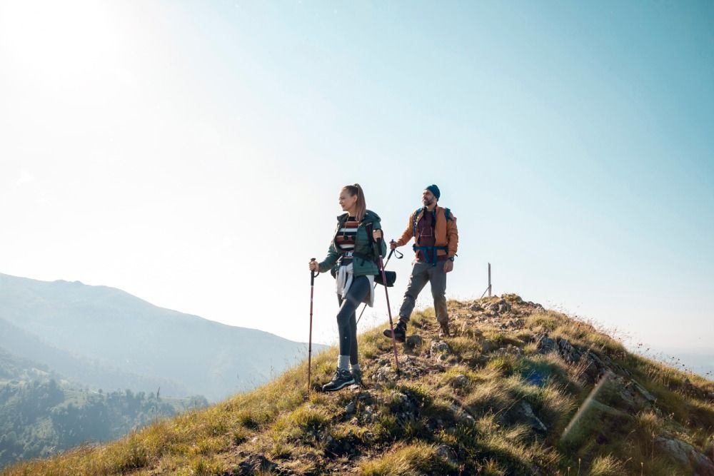 Two hikers with trekking poles standing on a grassy mountain ridge under a bright sky