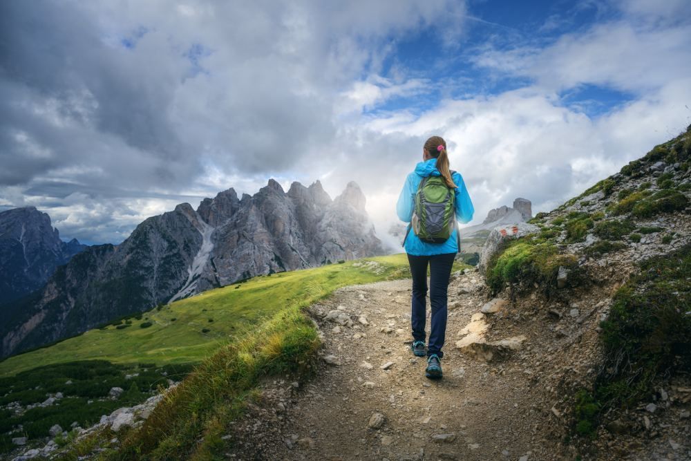 Hiker with blue backpack walking a mountain trail under a cloudy sky