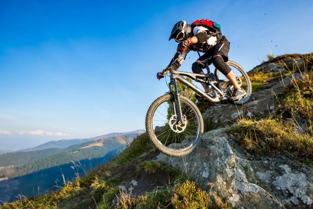 Mountain biker riding down a rocky trail on a sunlit hillside with blue sky and mountains in the background
