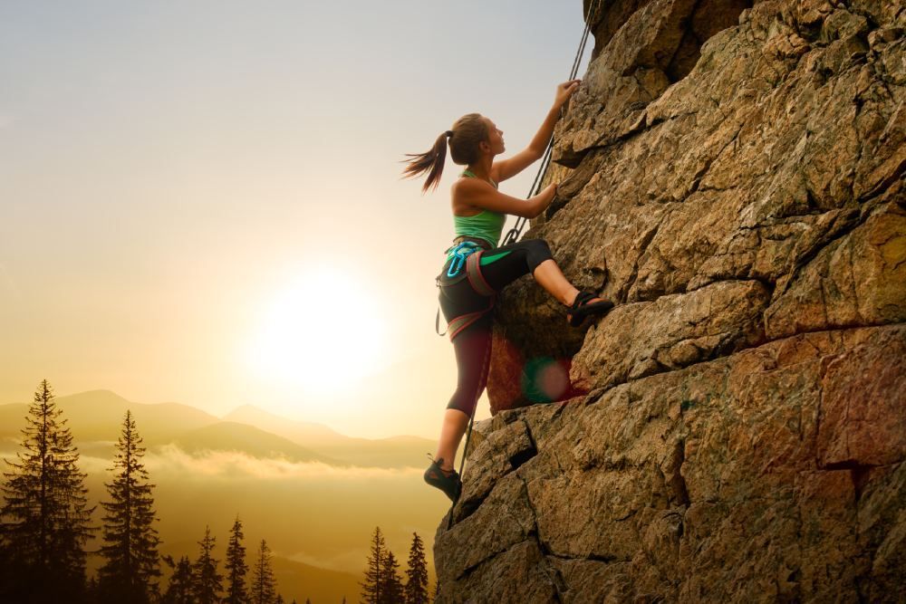 Two climbers scaling a rocky cliff at sunset, with a forest and glowing sky below
