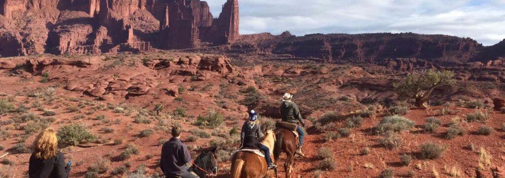 Cowboys on horseback crossing a red-rock desert beneath towering cliffs