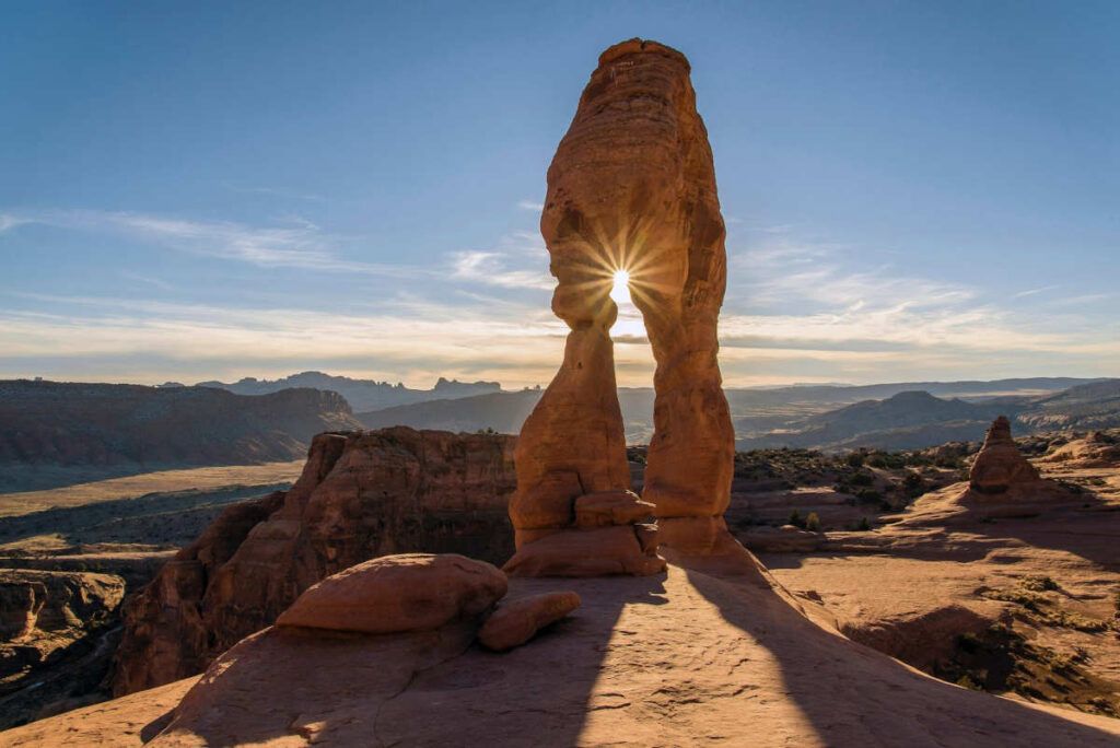 Sunlit sandstone arch in a desert landscape at sunrise, casting long shadows over distant mesas.